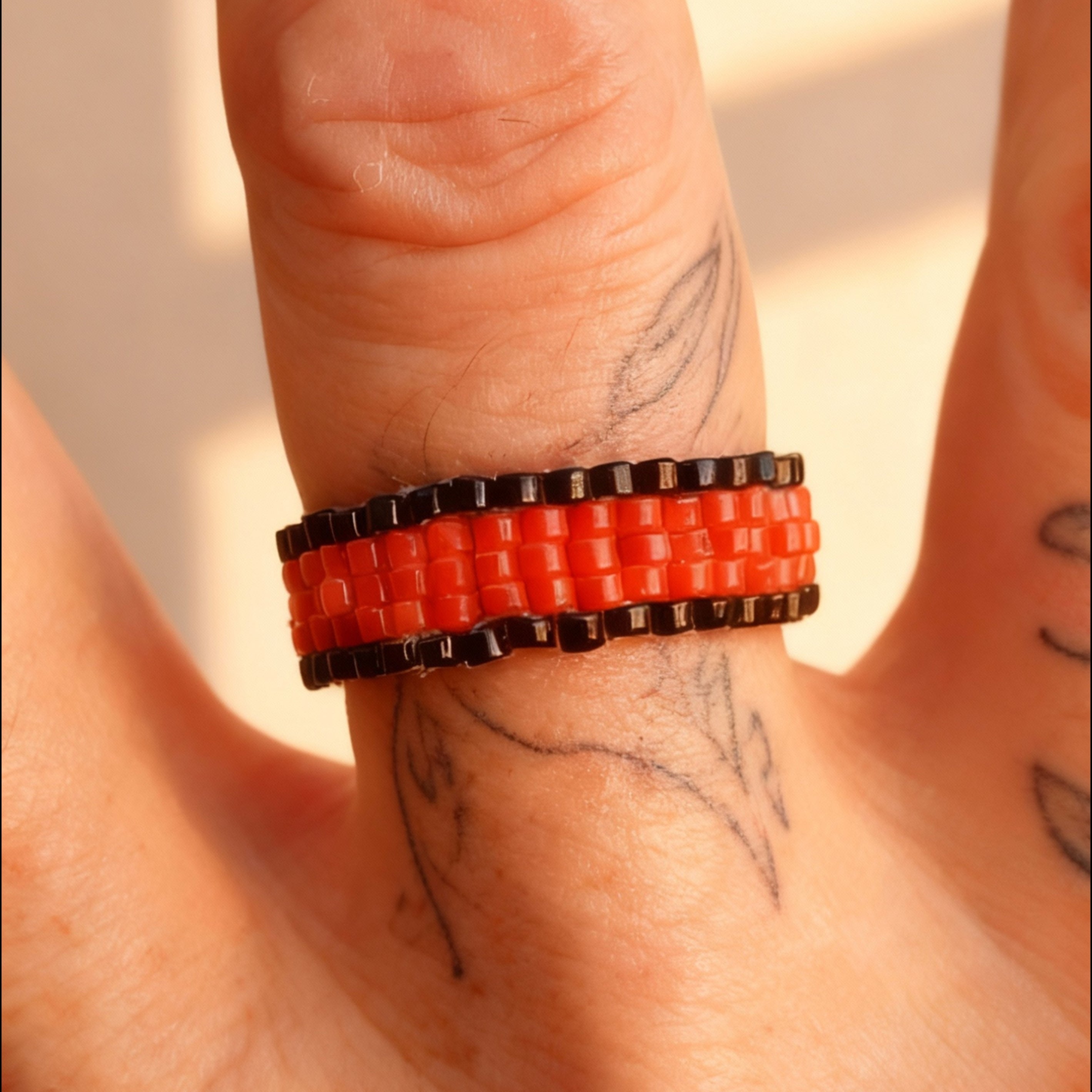 Close-up of a hand wearing a red and black beaded ring with tattoos on the fingers.